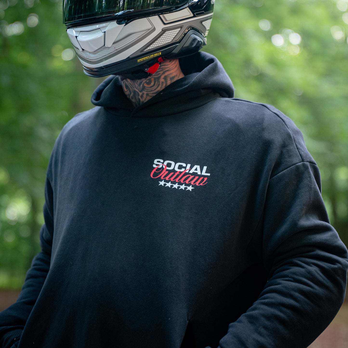 Person wearing a black hoodie with 'Social Outlaw' text, helmet, and tattooed neck outdoors.