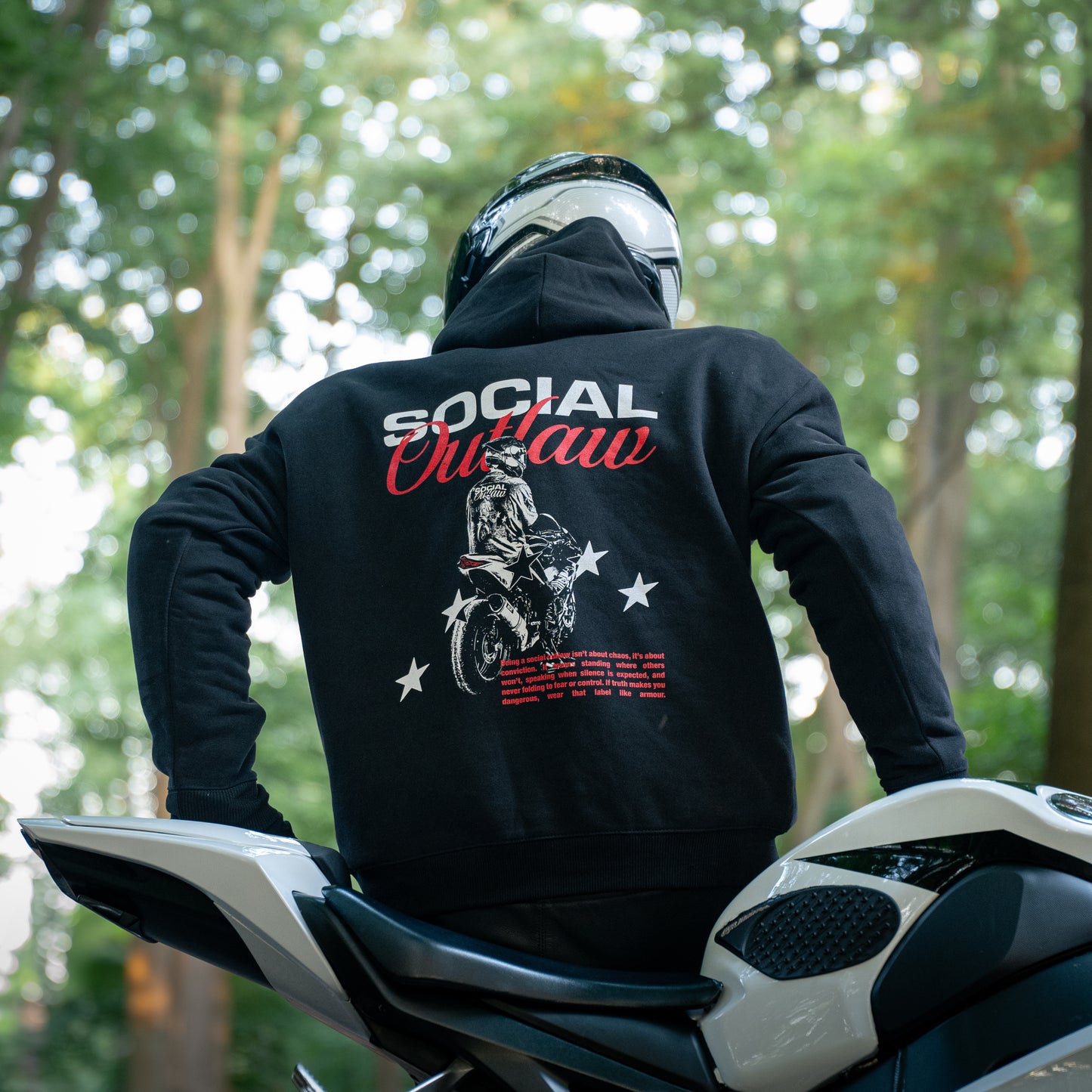 Person wearing a black hoodie with 'Social Outlaw' design on a motorcycle in a forest setting