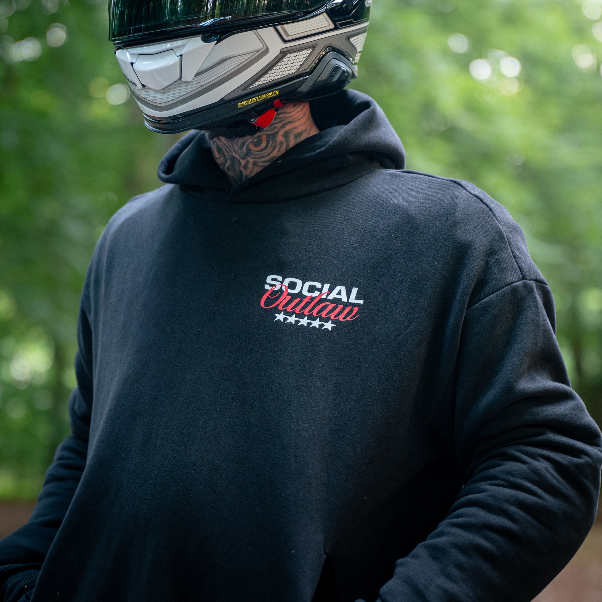 Person wearing a black hoodie with 'Social Outlaw' text, helmet, and tattooed neck outdoors.