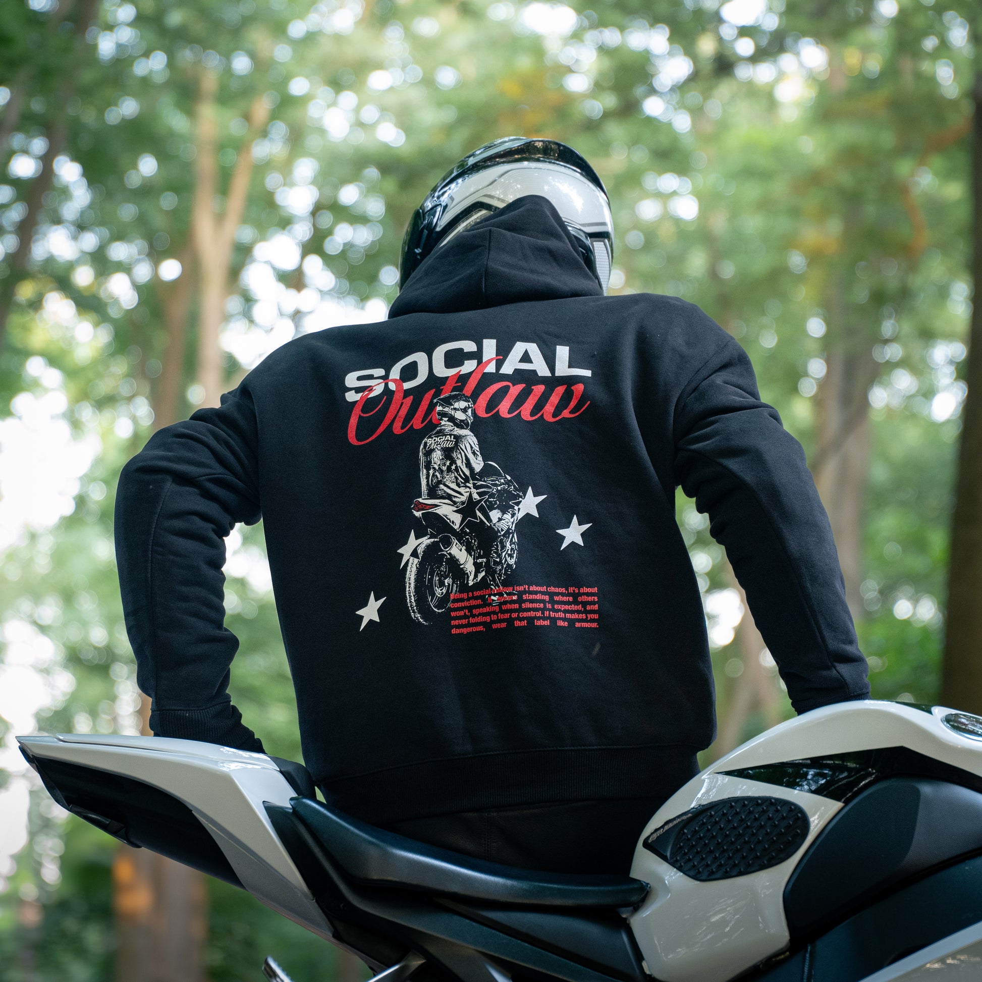 Person wearing a black hoodie with 'Social Outlaw' design on a motorcycle in a forest setting